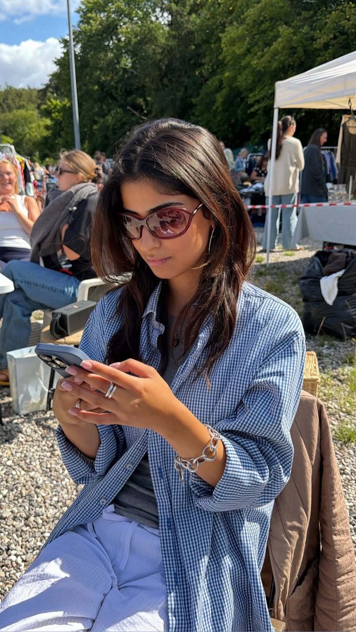Noor Aslani at an outdoor flea market in a blue gingham shirt and sunglasses, looking at her phone