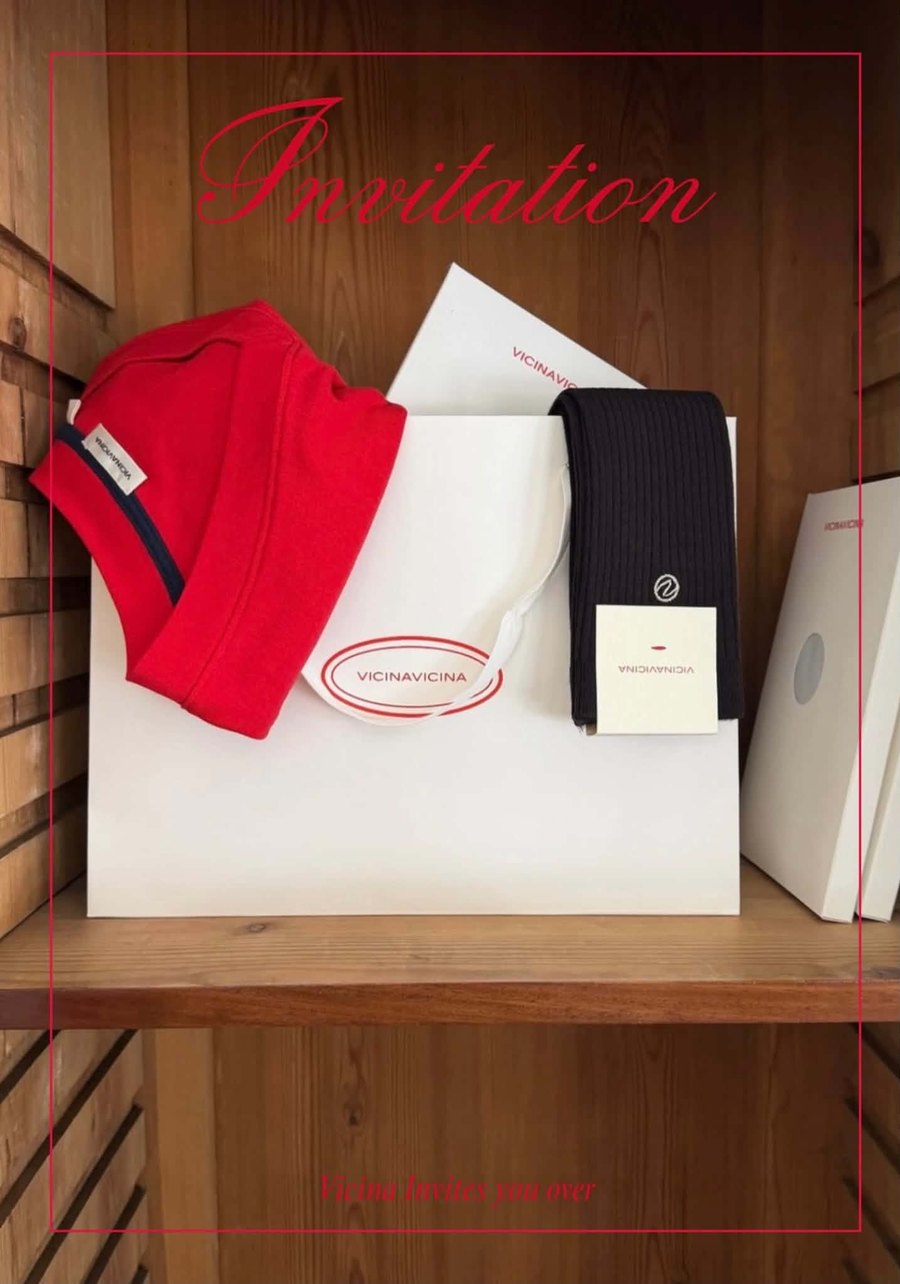 A VicinaVicina invitation styled with a red knit, navy socks and a branded paper bag on a wooden shelf